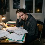 Un jeune homme de 26 ans fatigué et dépassé, se tenant la tête dans les mains à un bureau encombré de gros livres et de cours papier désordonnés