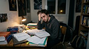 Un jeune homme de 26 ans fatigué et dépassé, se tenant la tête dans les mains à un bureau encombré de gros livres et de cours papier désordonnés
